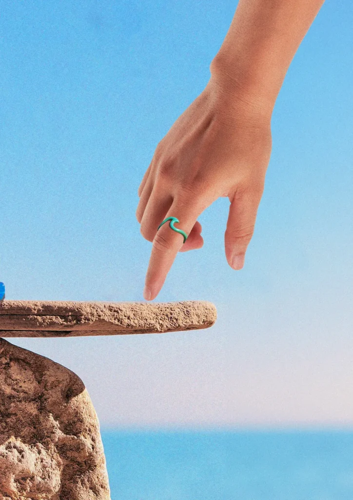 Mano con un anillo Wave turquesa sobre una roca, con el mar de fondo